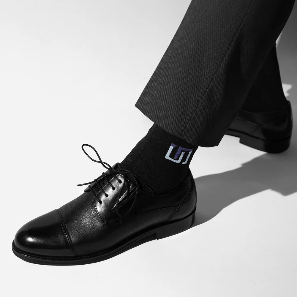 close-up of a person wearing black dress shoes and black wudhu socks with logo, paired with dark trousers